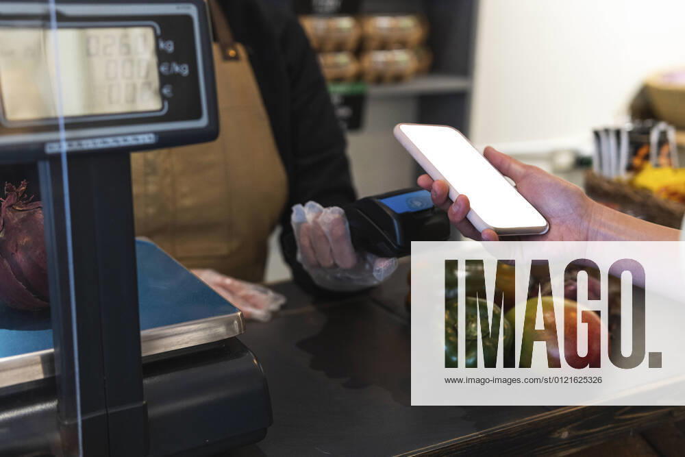 IMAGO Creative: Business, Inflation und Lieferketten Woman holding smart phone over credit card reader while paying at grocery store model released Symbolfoto property released PNAF01593