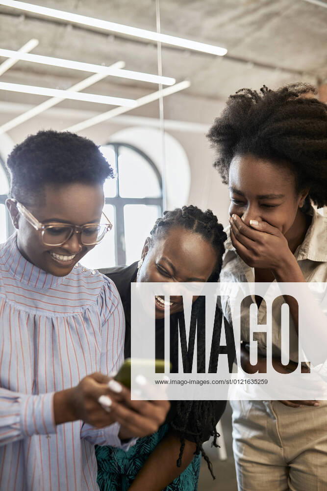 Happy female professional using mobile phone by female coworkers laughing at coworking office model released Symbolfoto property released ZEDF04145