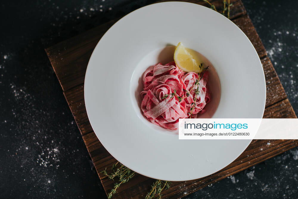 Top view of appetizing vegan pink beetroot pasta with lemon slice and thyme sprig served on wooden board on black table