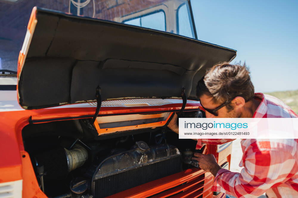 Side view of adult unshaven male traveler in casual clothes and sunglasses opening bonnet of retro van parked on sandy road during trip in nature, Model released, Property released