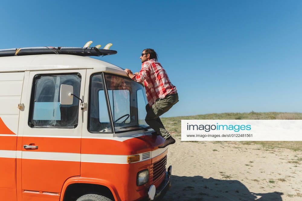 Young male traveler in casual clothes and sunglasses fixing surfboard on van roof against cloudless blue sky, Model released, Property released