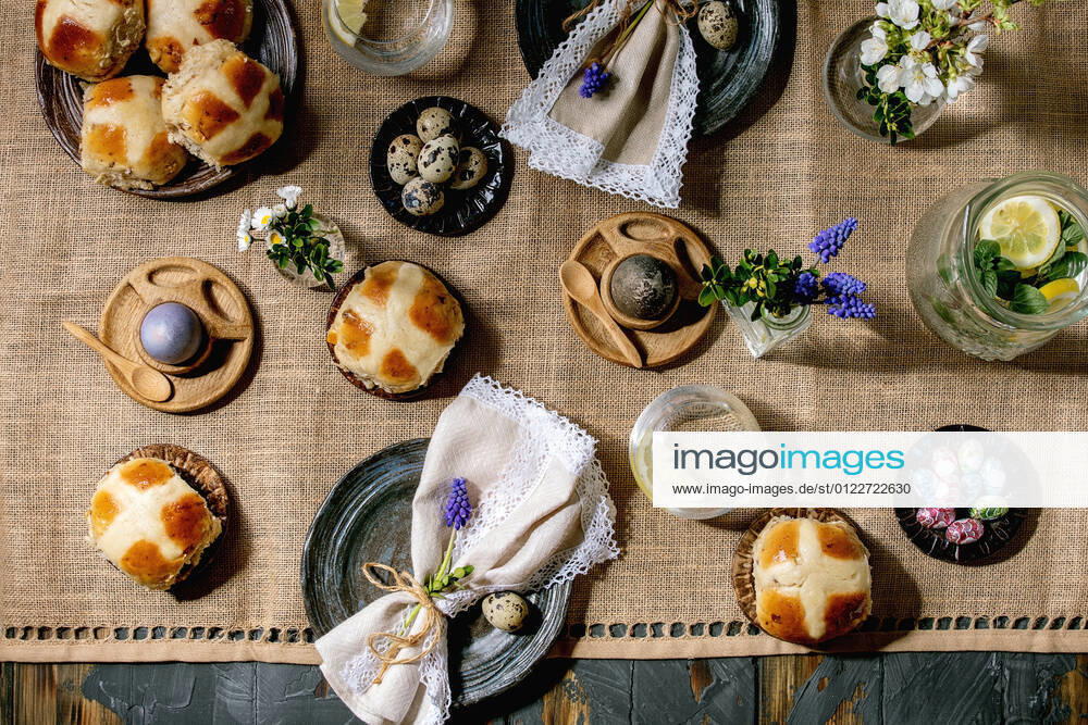 Easter table setting with colored and chocolate eggs, hot cross buns, bouquet flowers, empty ceramic plate with napkin, glass of lemonade drink on wooden table with textile tablecloth. Flat lay NatashaBreen REDA&CO 1028_29_NBR9686