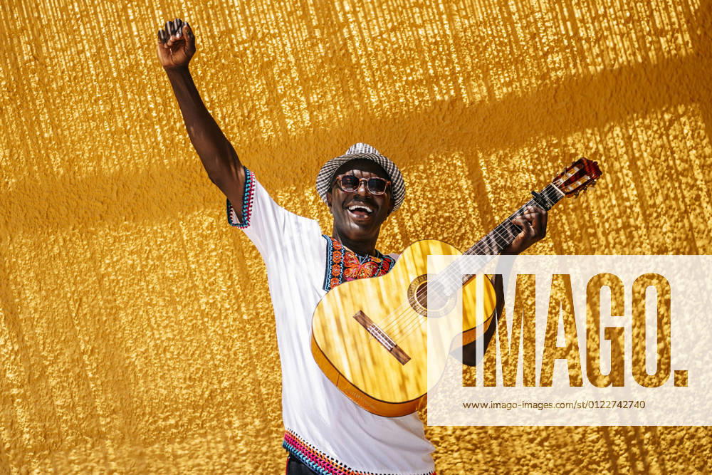 Cheerful man with hand raised carrying guitar in front of yellow wall during sunny day model released Symbolfoto OCMF02148