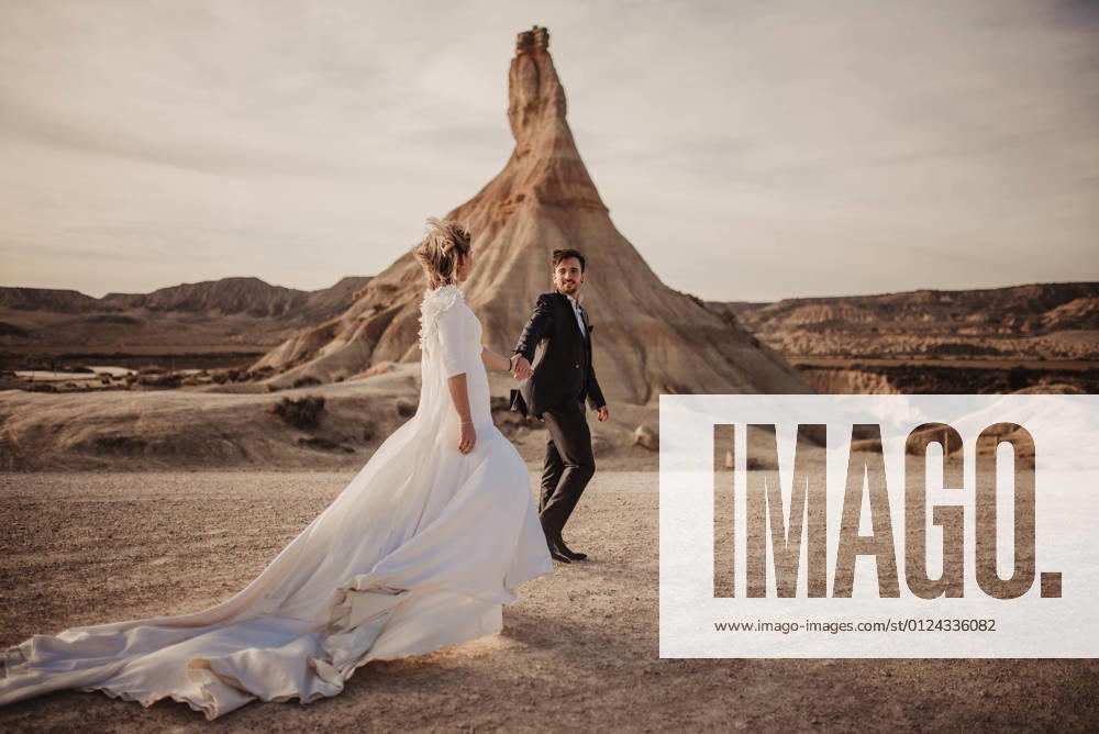 Happy groom and bride walking holding hands near mountain against cloudy sundown sky in Bardenas Reales Natural Park in Navarra, Spain, Model released