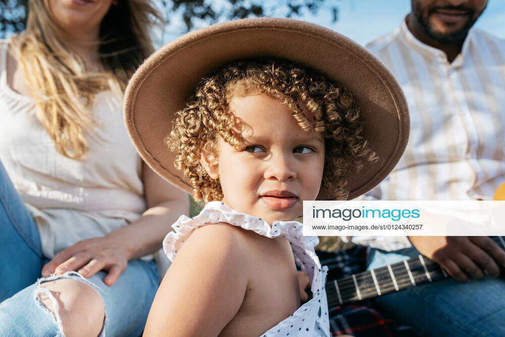 Delighted little girl with hat looking away sitting with multiracial family enjoying picnic together while playing guitar in nature
