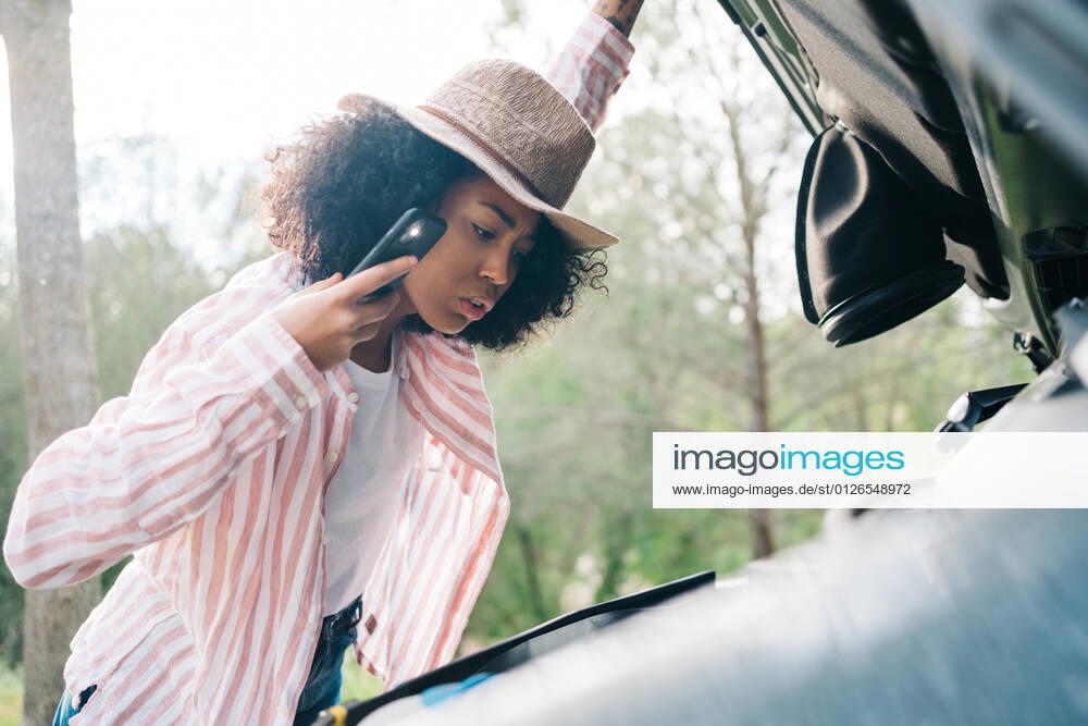 Side view of young African American female traveler holding smartphone with glowing flashlight while checking car engine as having problem during road trip through nature, Model released JakeJakab_CampingInTheNature_40.jpg