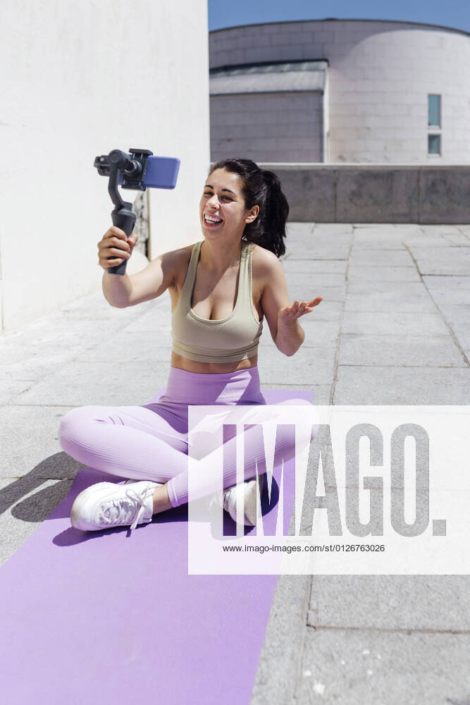 Smiling female athlete vlogging through smart phone while sitting on exercise mat during sunny day model released Symbolfoto EGHF00026