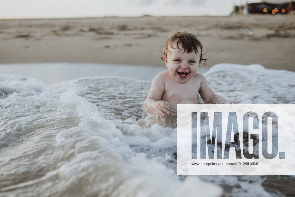 Male toddler laughing while sitting in water at beach during sunset model released Symbolfoto GMLF00754