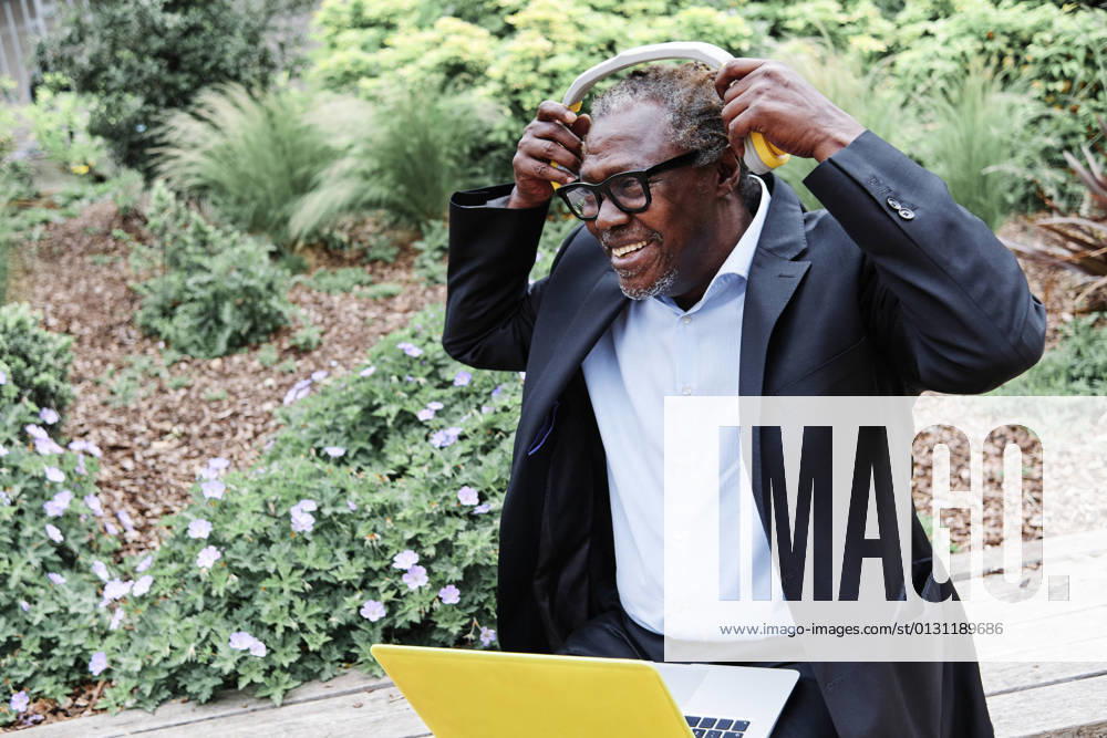 IMAGO Creative: Klimawandel - Zukunft und Innovation Smiling businessman wearing headphones while sitting with laptop on bench model released Symbolfoto ASGF00926