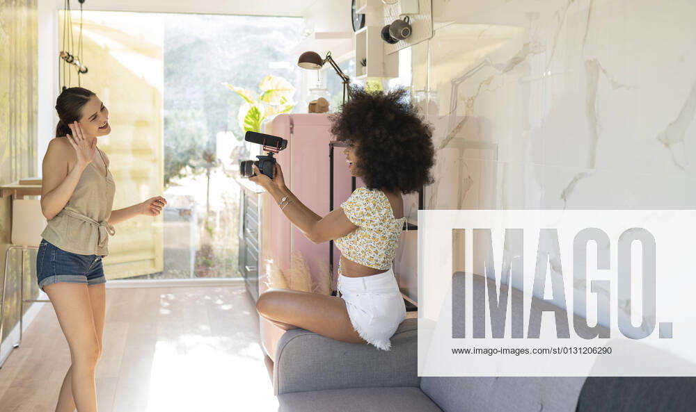 IMAGO Creative: Digitale Welt, Social Commerce Young woman waving in camera held by afro female friend while filming at home model released Symbolfoto property released JCCMF03221