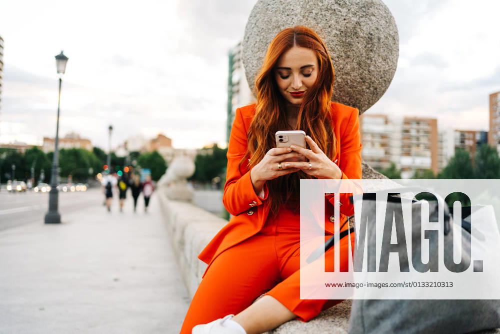 Happy stylish female with red hair and in vibrant orange suit sitting on stone border in city and messaging on cellphone JakeJakab_MariaPortraits_33.jpg