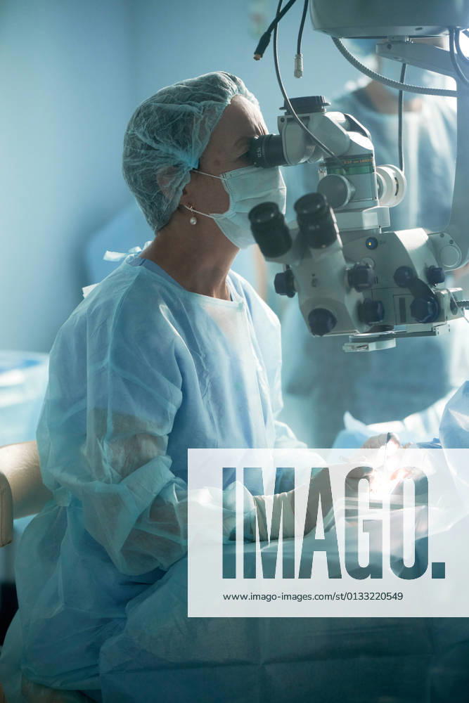 Adult female doctor in sterile mask and ornamental medical cap looking through surgical microscope against crop coworker in hospital, Model released SergeyMironov_Surgery_22.jpg