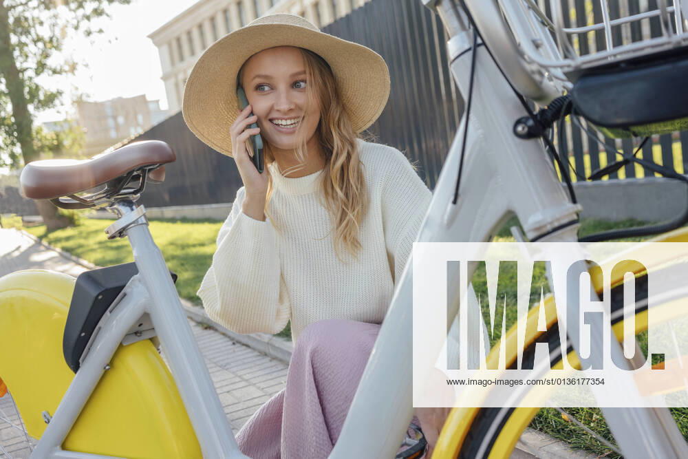 IMAGO Creative: Klimawandel - Zukunft und Innovation Smiling blond woman talking on smart phone while crouching by bicycle model released Symbolfoto VPIF04854