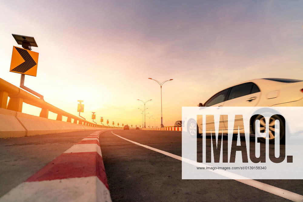 IMAGO Creative: Motorsport, Innovation und Nachhaltigkeit Motion blur of white car driving on curve concrete road with traffic sign. Road trip on summer vacation. Car drive on the street. Summer travel by car. Solar panel energy on yellow curve traffic sign