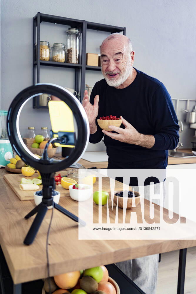 Senior male vlogger with fruit bowl filming through mobile phone at home model released Symbolfoto property released GIOF13790