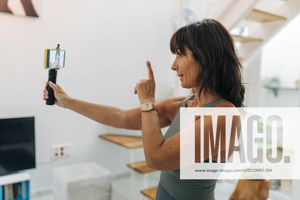 IMAGO Creative, Mainstream-Aktivismus Mature female influencer gesturing while filming video through smart phone at home model released Symbolfoto property released JRVF01877