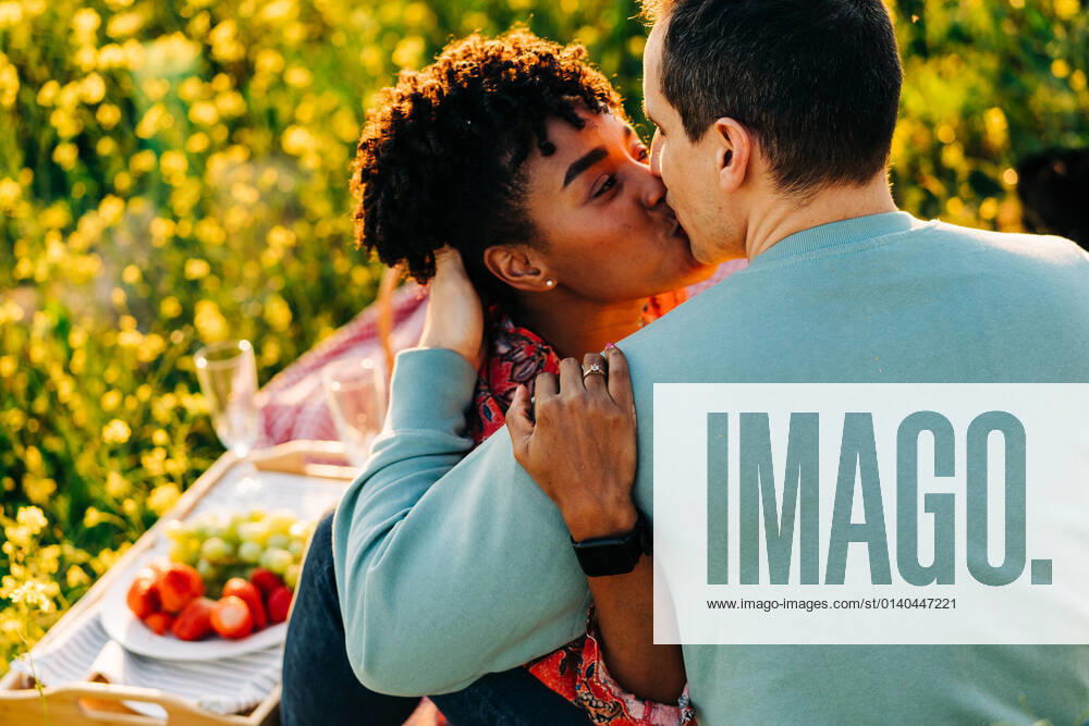 Young African American lady with curly hair embracing and kissing boyfriend with closed eyes during picnic on blooming meadow on sunny day, Model released JakeJakab_EliAndSergio_6.jpg