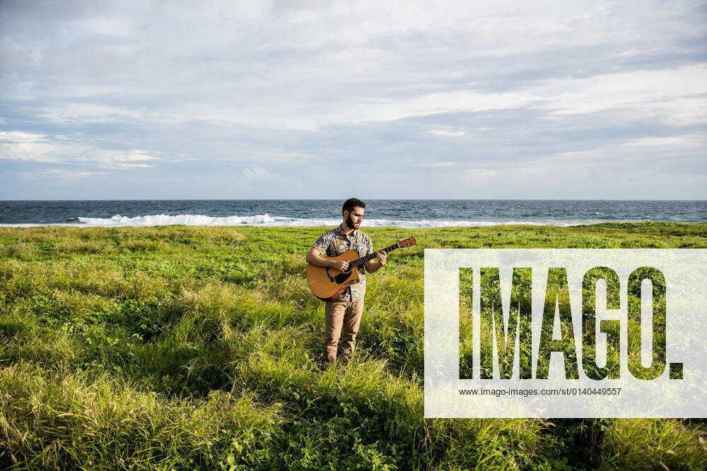 Calm man musician in casual clothes standing among green grass on coast of ocean and playing acoustic guitar in summertime in daylight, Model released LosMuertosCrew_MusiciansAtTheBeach_23.jpg