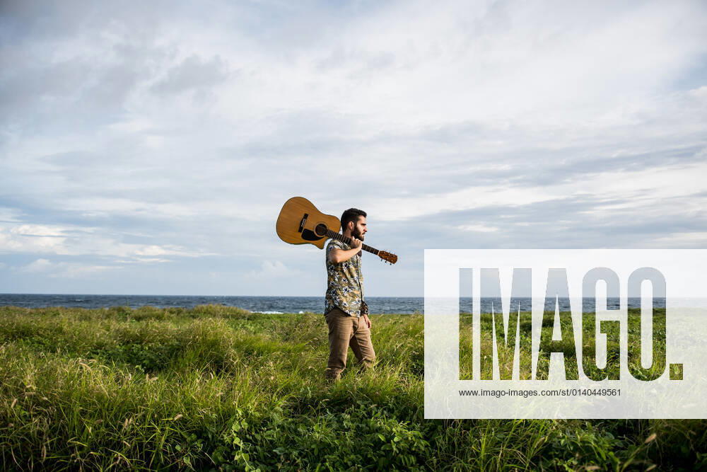Calm man musician in casual clothes standing with acoustic guitar on shoulder among green grass on coast of ocean in summertime in daylight, Model released LosMuertosCrew_MusiciansAtTheBeach_26.jpg