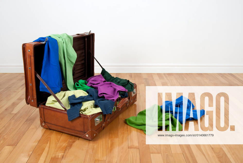 Leather suitcase on a wooden floor in an empty room, full of colorful clothing.