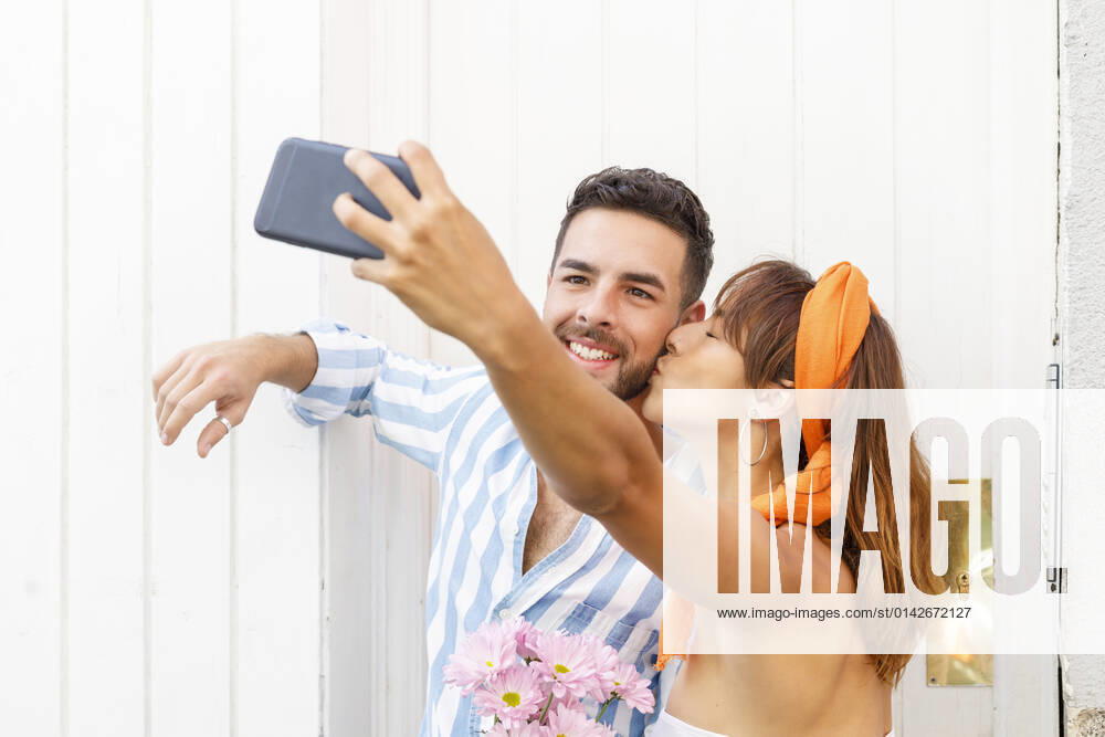 Girlfriend taking selfie through smart phone while kissing boyfriend by white wall model released, IFRF00810