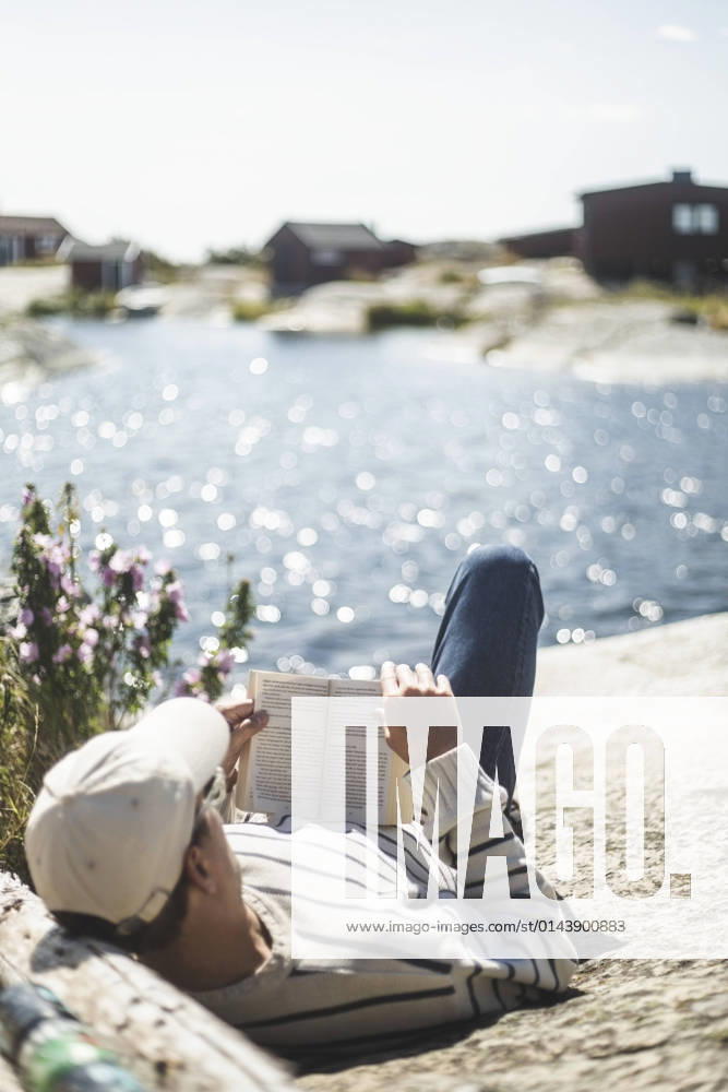 IMAGO Creative, Jahreszeit Sommer, Sommerferien Man reading book while lying down on rock during sunny day Sweden, Stockholm archipelago, model released