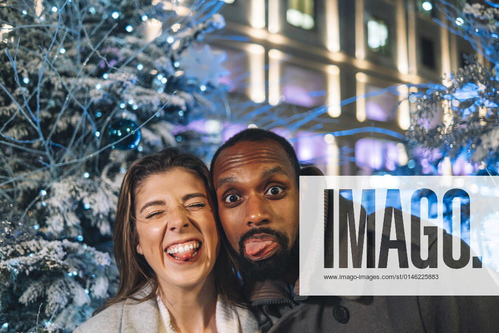Playful couple sticking out tongues near Christmas tree at night model released, ASGF01858