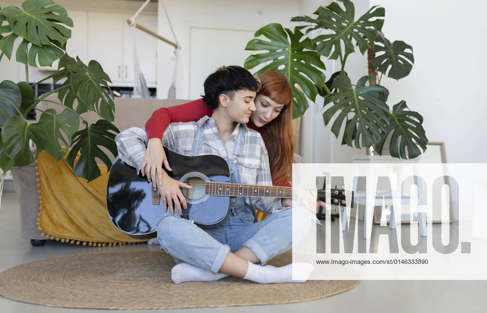 IMAGO Creative: Bildung, Musikerziehung Content lesbian hugging talented female partner playing on acoustic guitar on rug in light living room with green potted plants JoseCarlosIchiro_CoupleWomaenAtLoft_37.jpg
