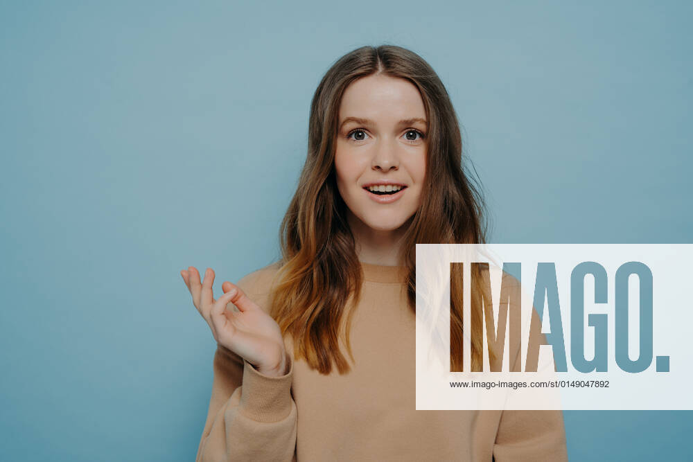 Pretty young girl showing amusement and surprise posing against blue wall Smiling teenage girl raising hand and looking ahead with amusement and surprise, brunette with wavy hair wearing casual light brown sweater posing against blue background