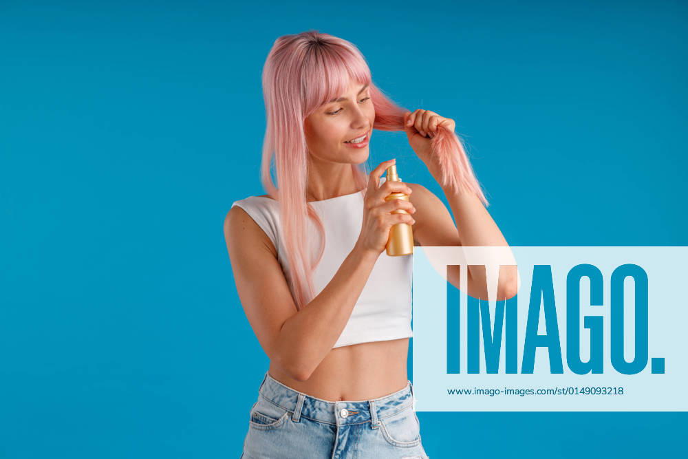 IMAGO Creative, Lifestyle, Barbiecore Caring young woman smiling and applying moisturizing spray to her smooth pink hair while standing isolated over blue studio background Caring young woman smiling and applying moisturizing spray to her smooth pink hair while standing isolated over blue studio background. Beauty products, hair care concept