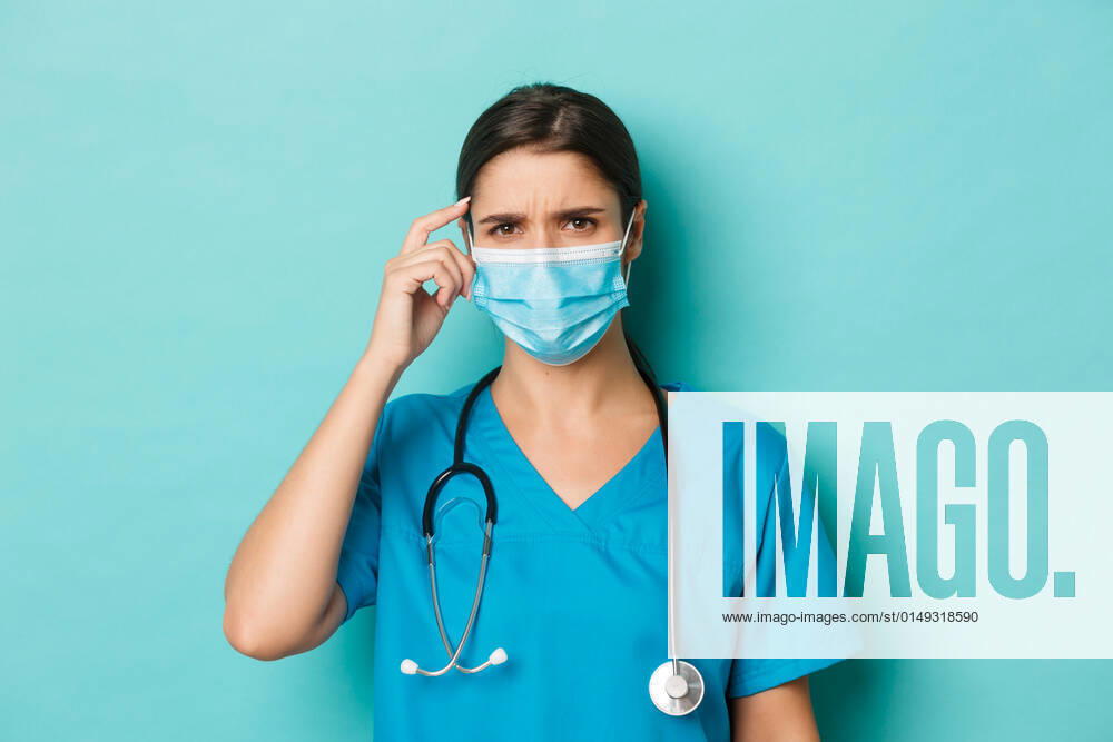 Concept of covid-19 and quarantine concept. Close-up of confused female doctor in medical mask and scrubs, frowning and looking perplexed, standing over blue background Concept of covid-19 and quarantine concept. Close-up of confused female doctor in medical mask and scrubs, frowning and looking perplexed, standing over blue background.