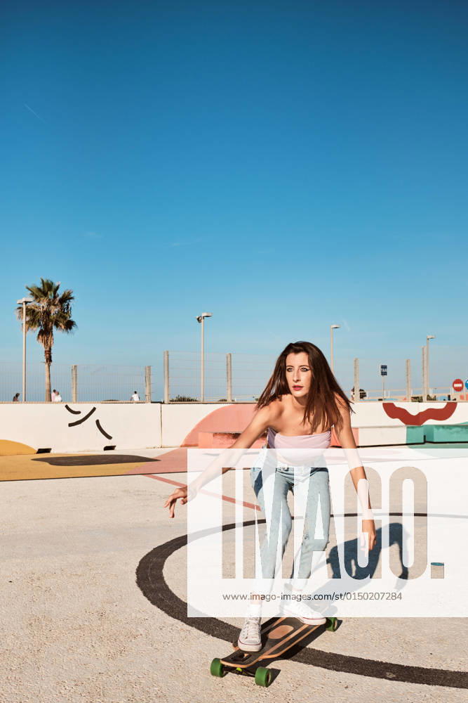 IMAGO Creative: Sommer, Sommertage im Stadtpark Full body of focused slim young female with long dark hair wearing top and jeans riding longboard in skate park in sunny summer day and looking away, Model released ManuelOrts_SkateYoungWoman_4.jpg