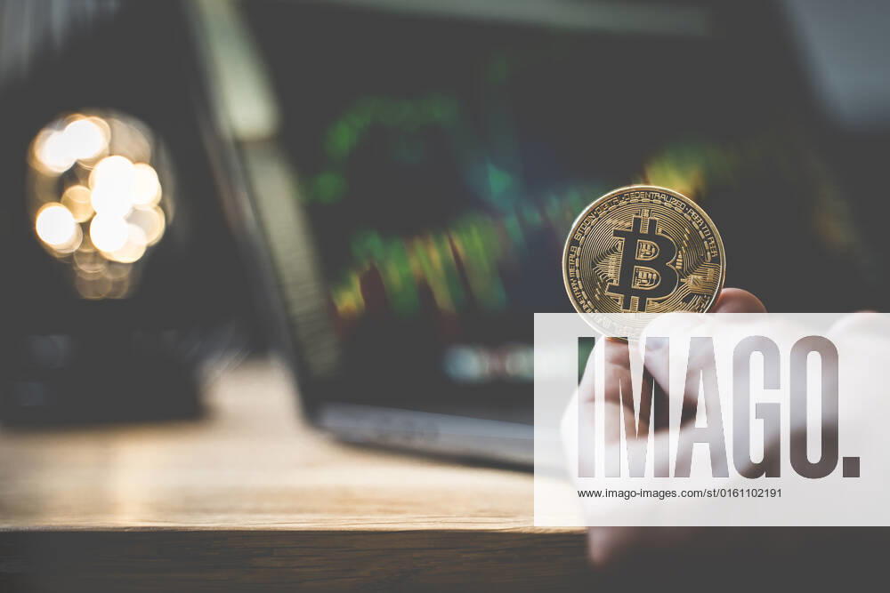 Hand holding a Bitcoin gold coin BTC cryptocurrency in front of a laptop computer, online trading on stock exchange with price chart