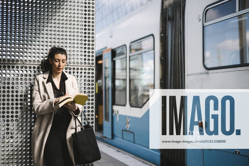 Businesswoman wearing long coat reading book while waiting at railroad station Sweden, Gothenburg, model released