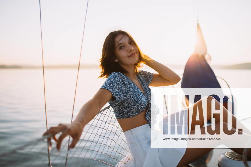 Side view of positive young stylish female traveler with dark hair in trendy crop top and skirt looking at camera while sitting on sailboat during cruise under sunset sky, Model released CamilleGirard_Sailboat_00029.jpg