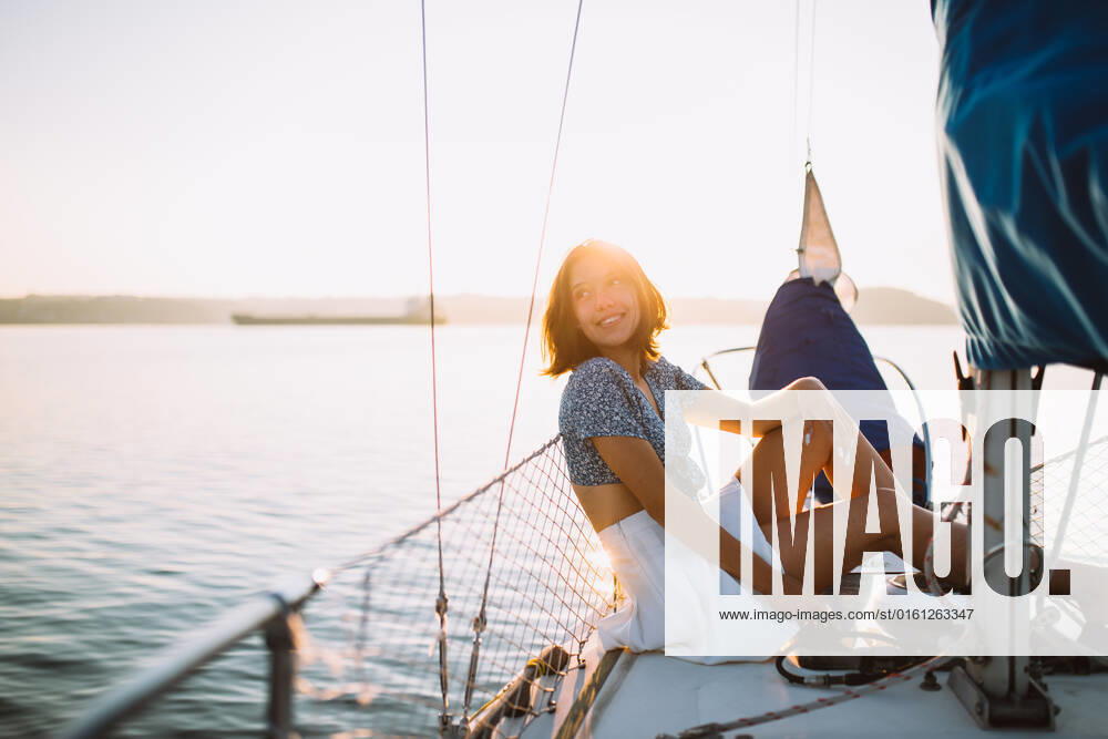 Side view of positive young stylish female traveler with dark hair in trendy crop top and skirt looking away while sitting on sailboat during cruise under sunset sky, Model released CamilleGirard_Sailboat_00030.jpg