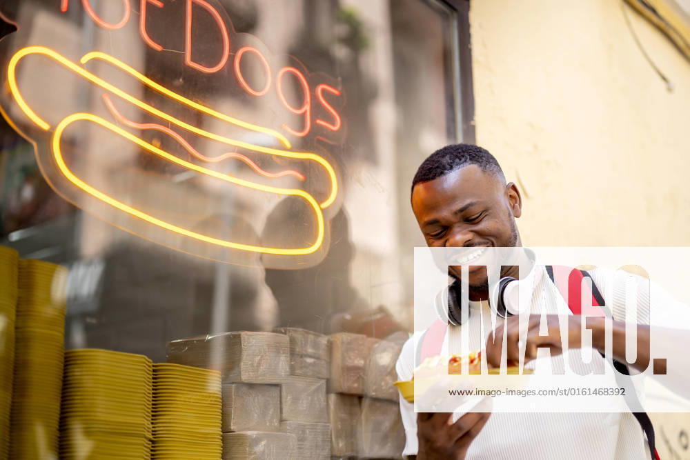 IMAGO Creative, Urban Lifestyle Happy young man eating hot dog leaning on restaurant window model released, Symbolfoto property released, OCMF02415