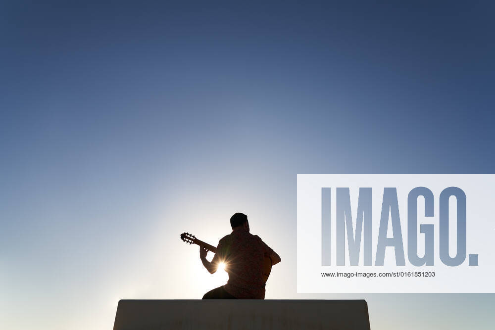 Back view silhouette of unrecognizable bearded musician playing song on acoustic guitar while sitting on border against blue cloudless sky on sunny day in city, Model released JavierBermudez_RubenGuitarrist_13.jpg