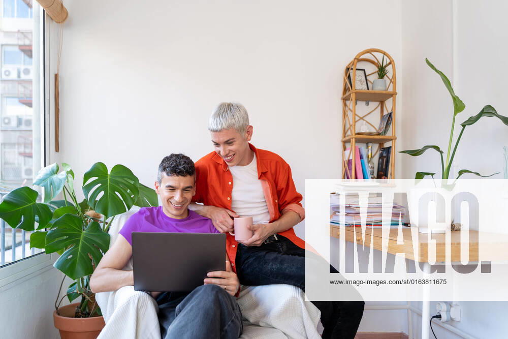 Smiling gay couple using laptop sitting on armchair at home, Model released JordiBataller_QueerReality_129.jpg