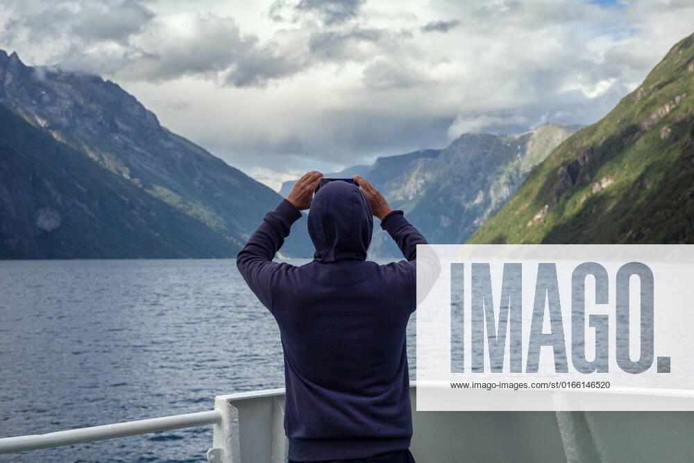 Ferry ride on Geirangerfjord Tourist stands at the stern of a ship and takes photos of the breathtaking mountain scenery with the fjord with his cell phone, model released Norway