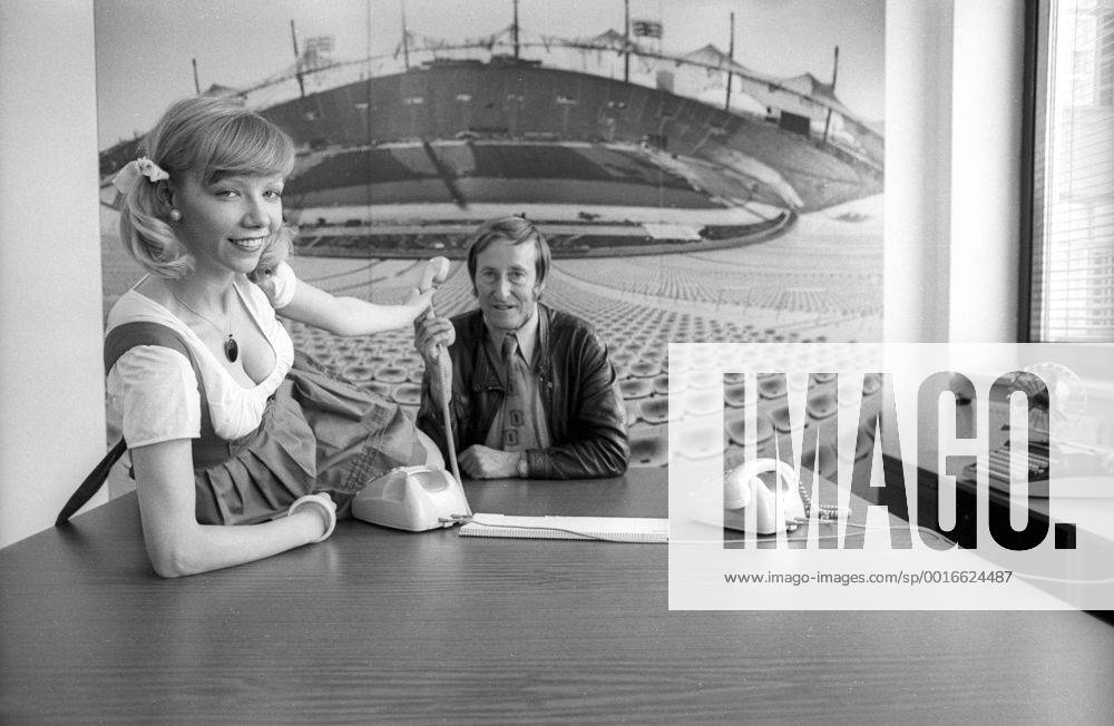 Rudi Gutendorf (BR Deutschland) sitzt vor einem Bild des Münchner Olympiastadions an einem Tisch, auf dem sich eine Frau im Dirndl rekelt