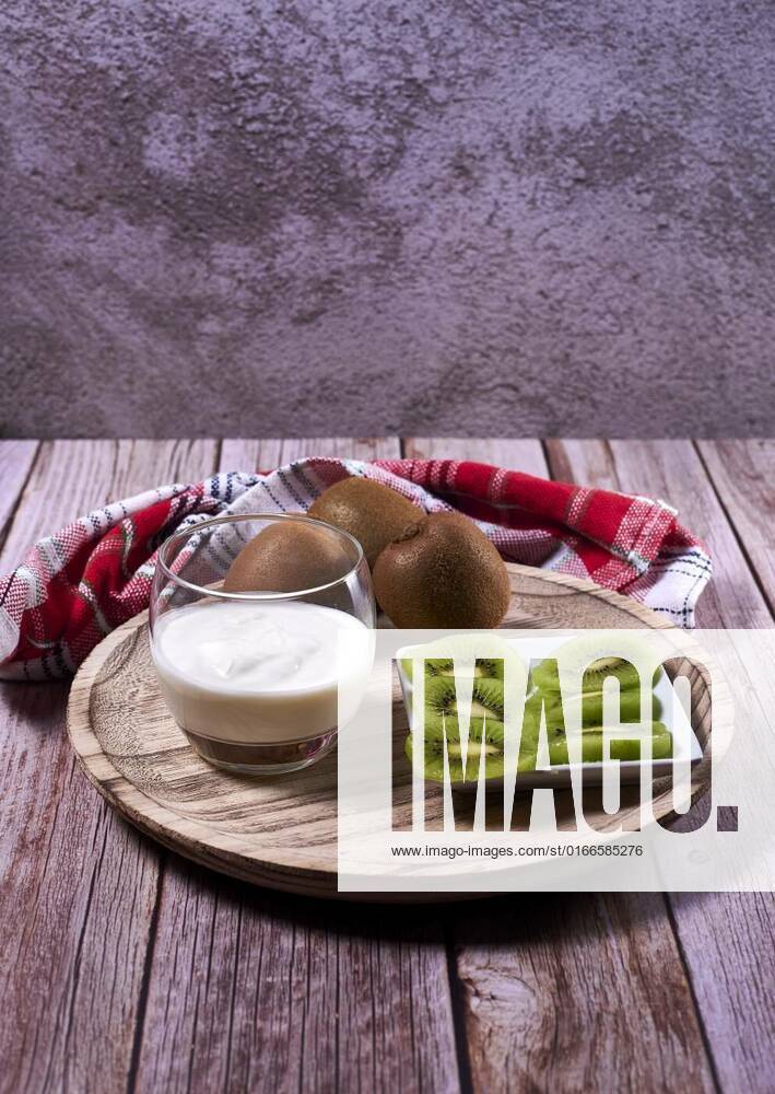 Glass with yoghurt and broken kiwis on a wooden plate, Glass with yoghurt and broken kiwis on a wooden plate. Wooden floor, whole kiwis, white and red kitchen towel, stone wall., 27.02.2021,