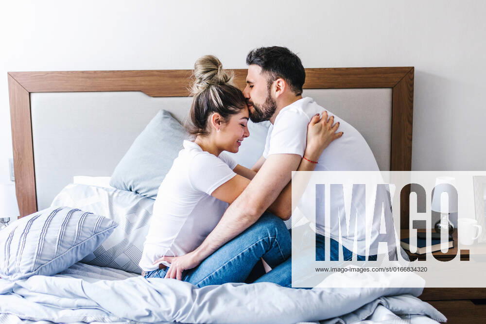 Side view of gentle Latin man kissing girlfriend on forehead while embracing on soft bed in bedroom, Model released, Property released MarcosCastillo_LatinYoungCouple_41