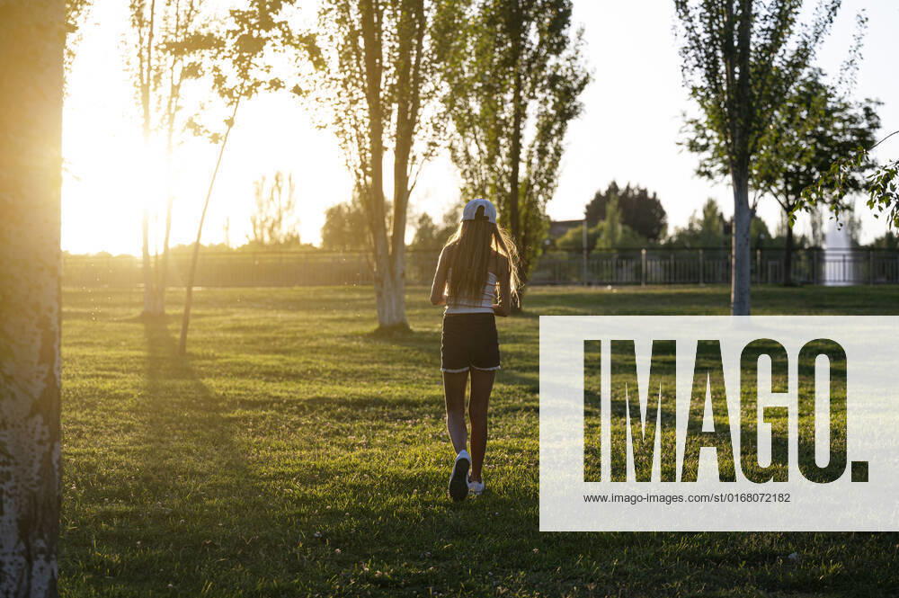 IMAGO Creative: Sommer, Sommertage im Stadtpark Full body back view of anonymous girl in cap strolling on grassy lawn in park with trees on summer evening, Model released JoseLuisCarrascosa_ID441_16966_120