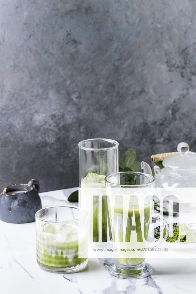 Matcha iced latte Matcha green tea iced latte or cocktail in three different glasses with ice cubes, matcha powder and jug of milk on white marble table, decorated by green branches. Grey wall at background