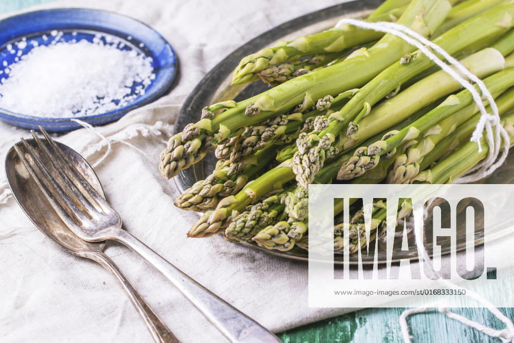 Bunch of young green asparagus on vintage tray served with ceramic plate of sea salt and silver cutlery over green wooden table.