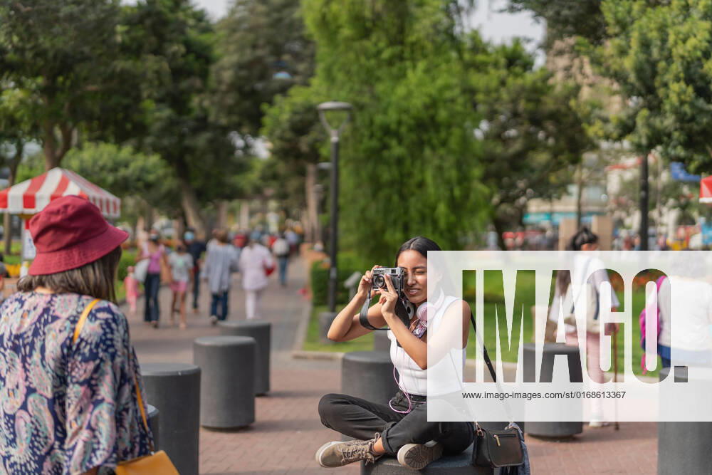 IMAGO Creative: Sommer, Sommertage im Stadtpark Peruvian female taking picture of anonymous female friend on photo camera while sitting on crowded street of city with trees, Model released GuillermoSpelucin_SaraiAndPaulaInLima_12