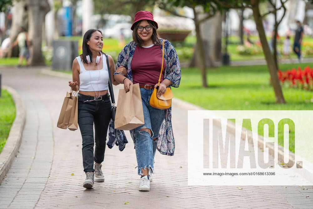 Full body of positive Peruvian female friends in casual wear with paper bags strolling on paved walkway in park on summer day, Model released GuillermoSpelucin_SaraiAndPaulaInLima_9
