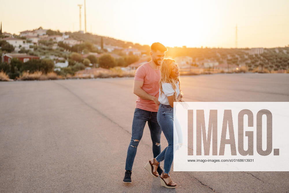 Cheerful man and excited woman looking at each other and laughing while walking dancing holding hands along asphalt road against blurred town on hillside in evening, Model released GustavoPozo_IsaAndKike_99