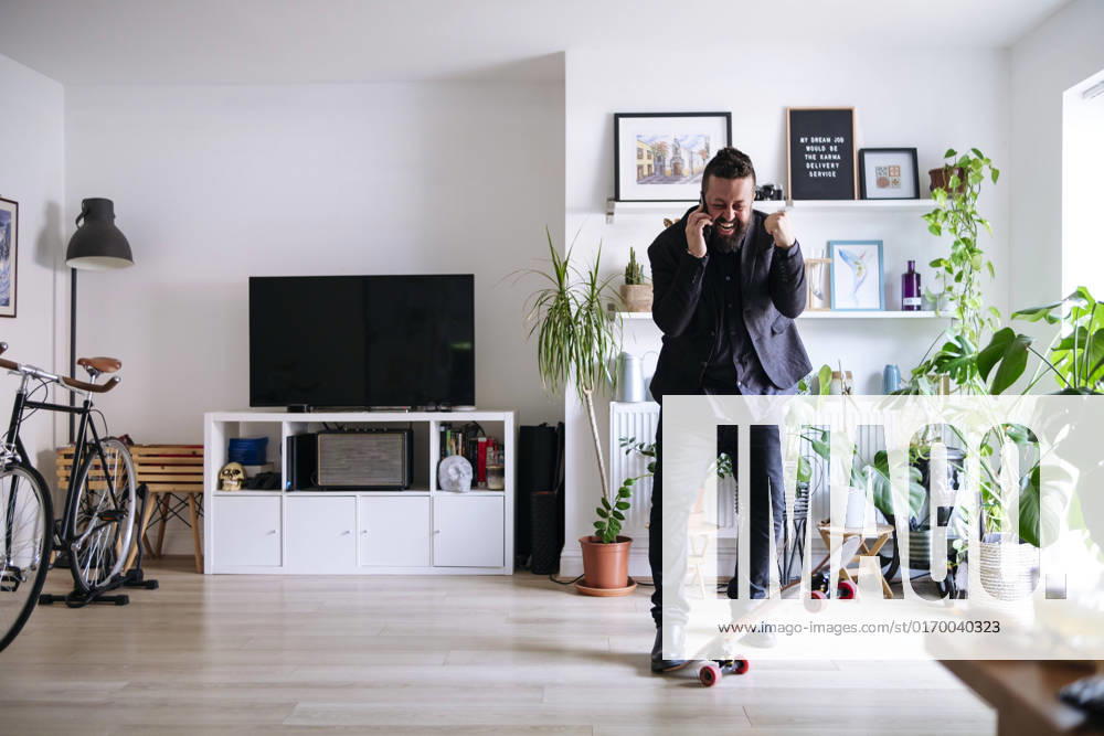IMAGO Creative: Business, Innovative Technologie-Lösungen Cheerful businessman with skateboard talking on smart phone and clenching fist at home model released, Symbolfoto property released, ASGF02709